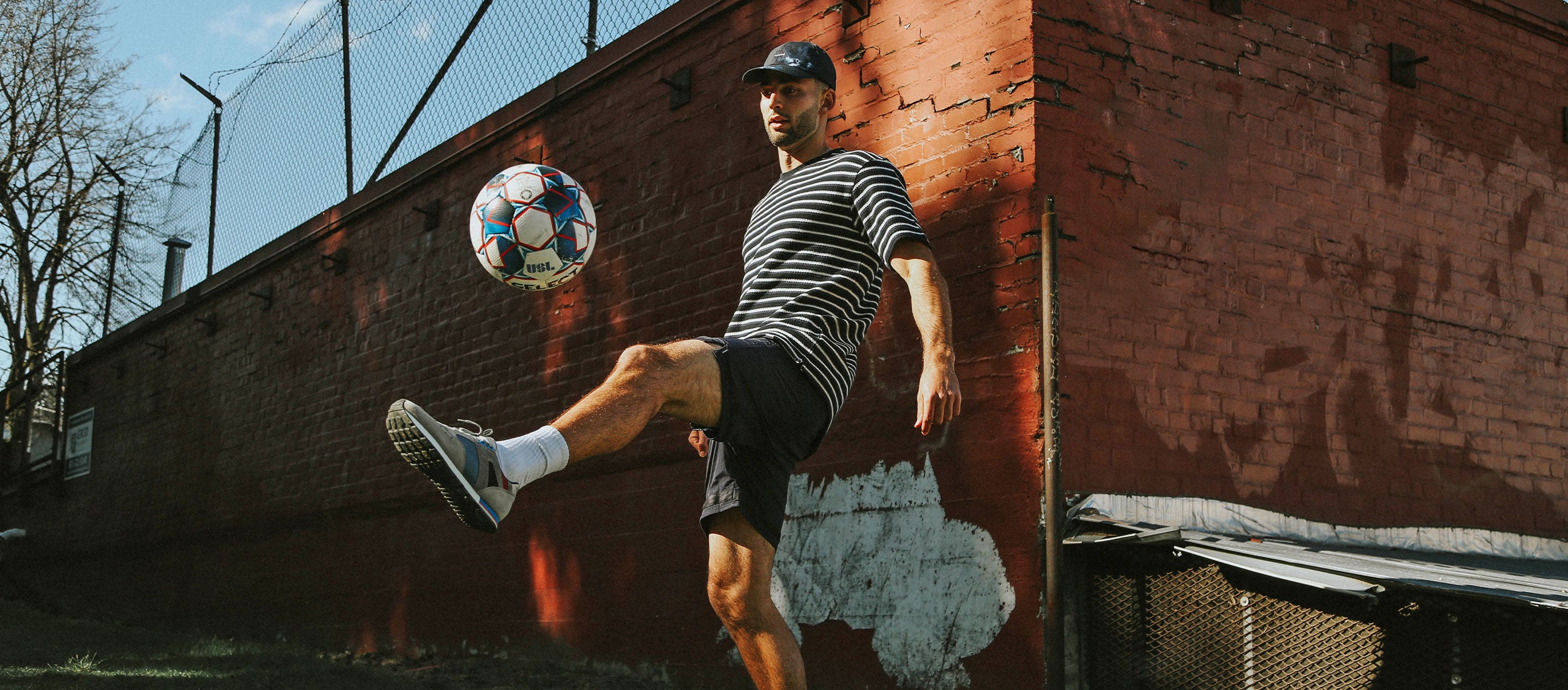 A young man juggling a football, or soccer ball, in front of a red brick wall.