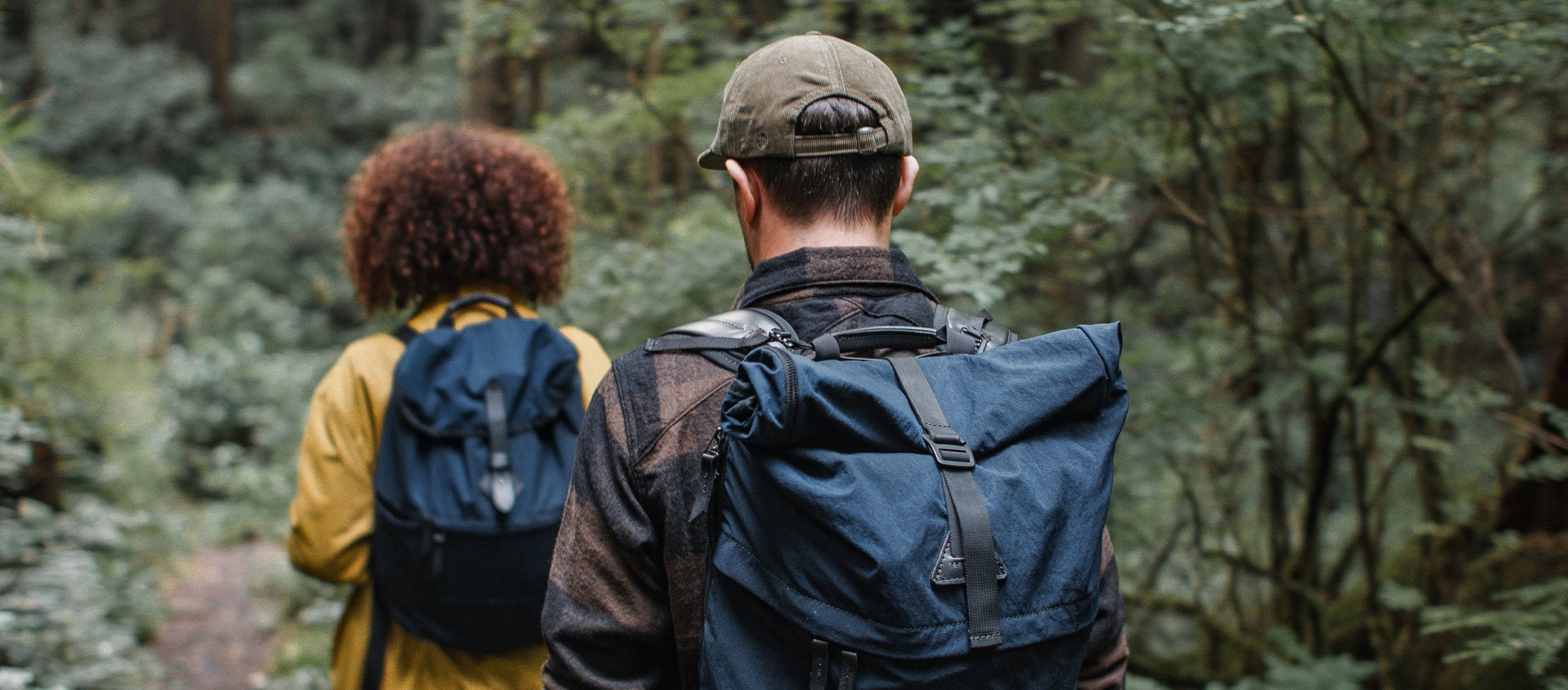 Two hikers on a path in a dense forest wearing navy blue backpacks from Tanner Goods.